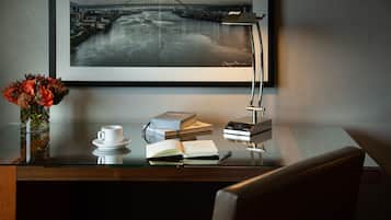 A desk with a glass top, a vase of flowers, a cup, a book, a lamp, and a framed picture on the wall.