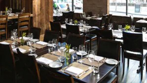 Un interior de restaurante con mesas y sillas de madera, ventanas amplias y lámparas colgantes.
