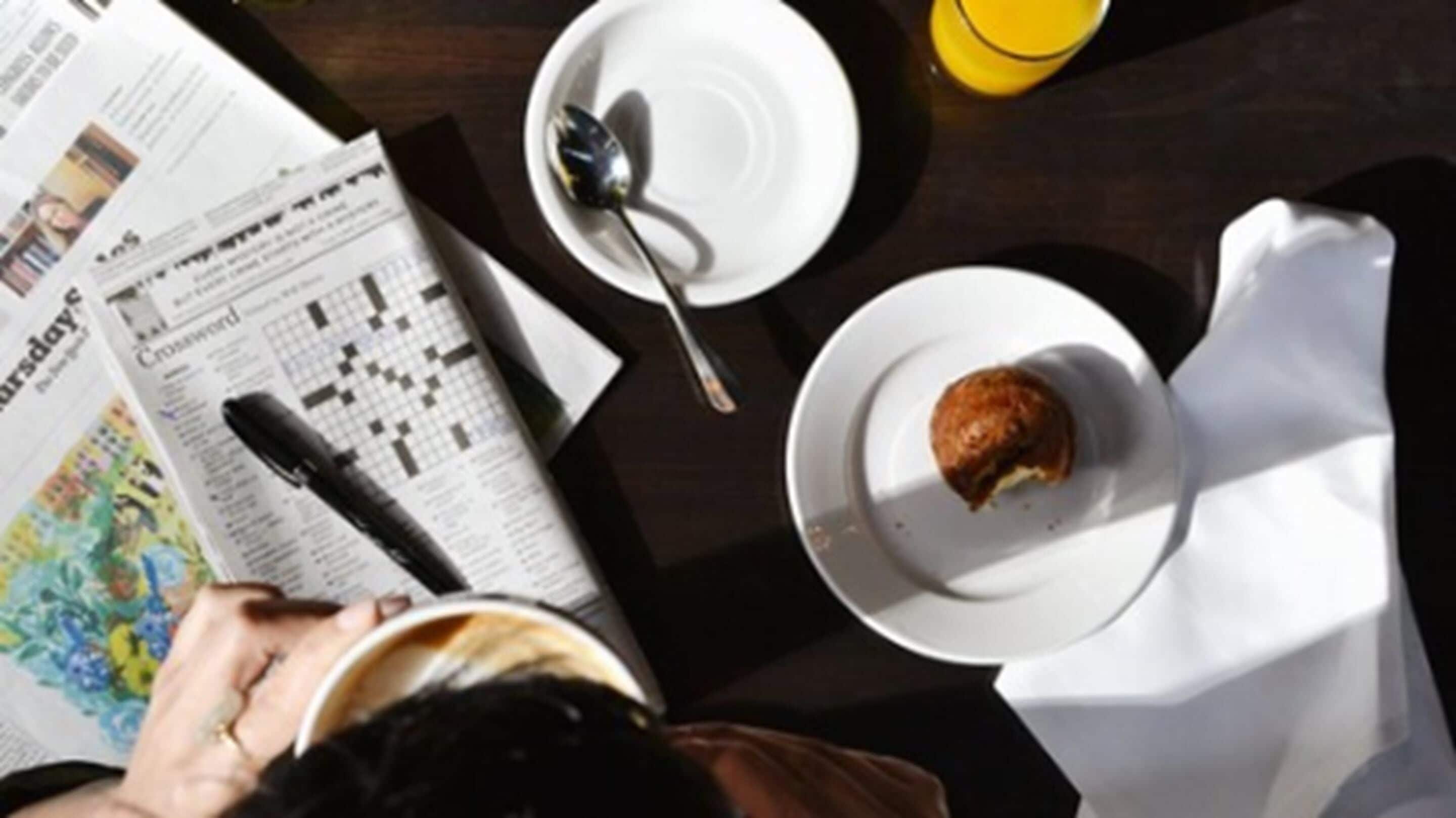 A breakfast setting with a muffin on a plate, a cup of coffee, a spoon, and a newspaper.