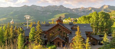 A log cabin with a large front porch, surrounded by trees and mountains in the background.