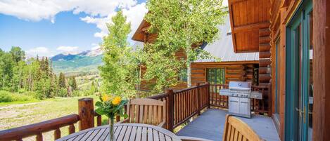 A wooden deck with a table and chairs, a barbecue grill, and a view of mountains and trees.