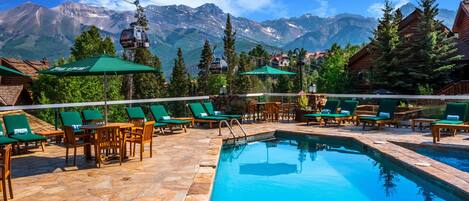 A pool area with lounge chairs and umbrellas, surrounded by mountains and wooden cabins.