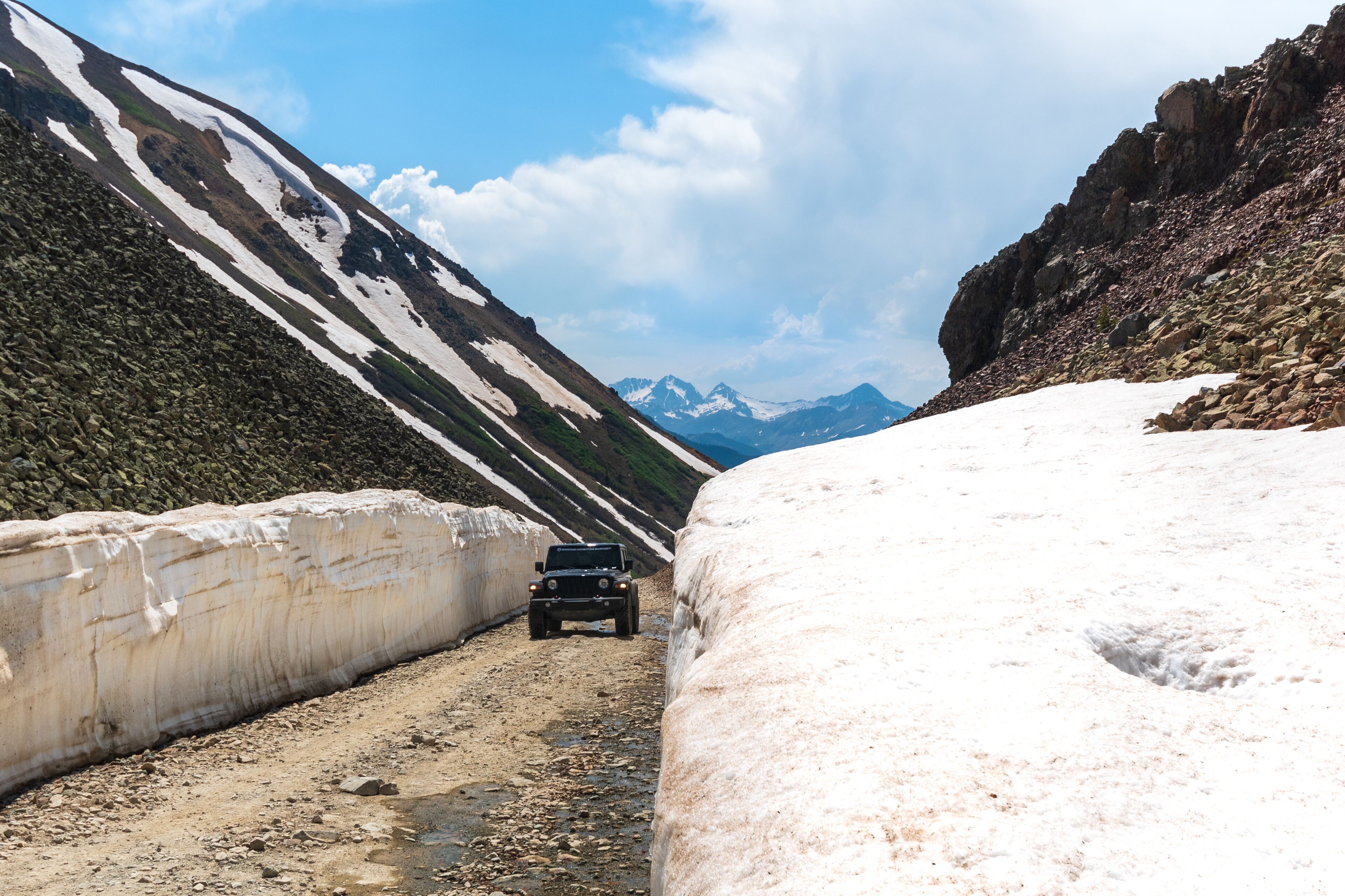 Een ruige bergweg met een voertuig, besneeuwde hellingen en verre bergtoppen.