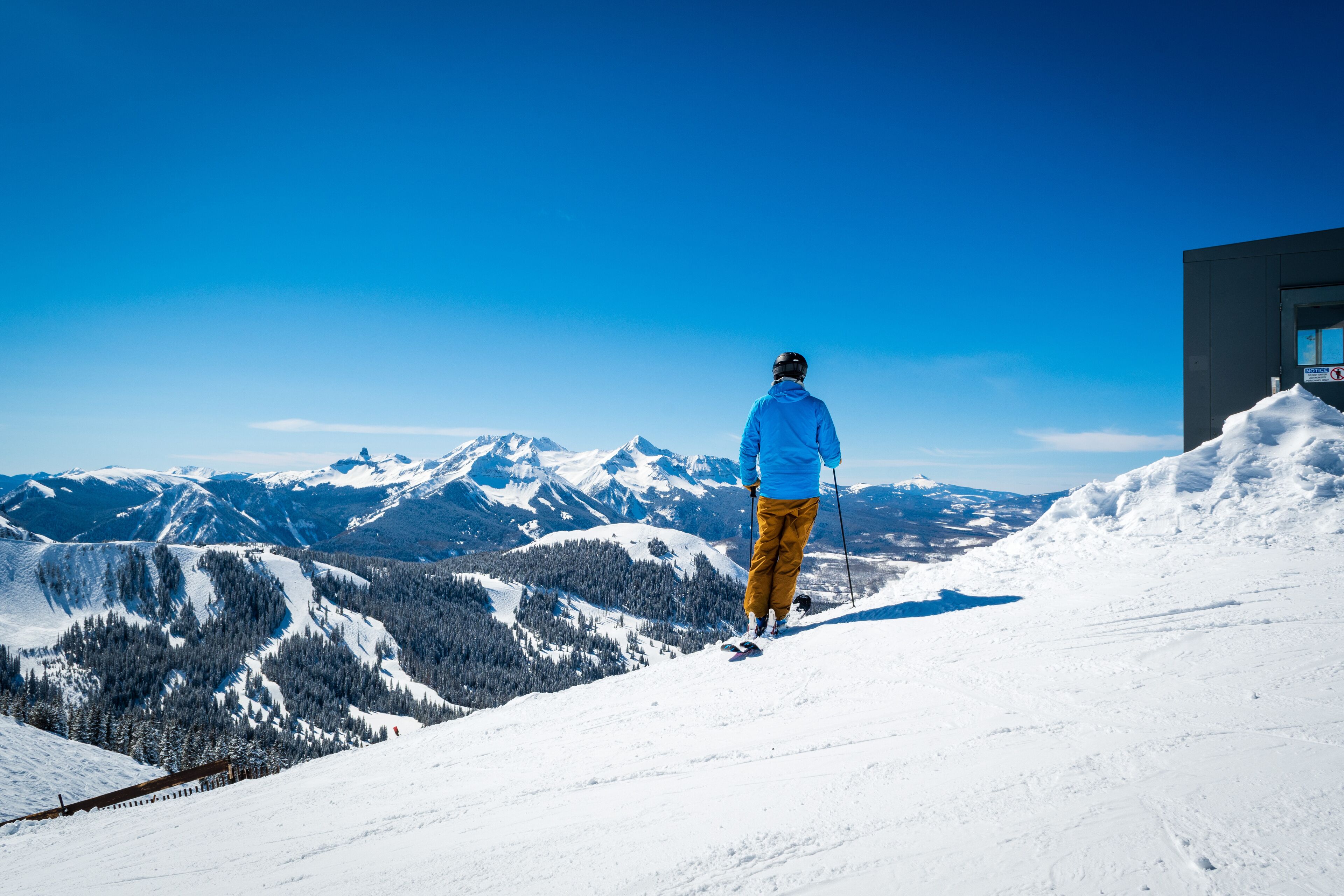 A person skiing on a snowy mountain slope with a clear blue sky and snow-covered peaks in the background.