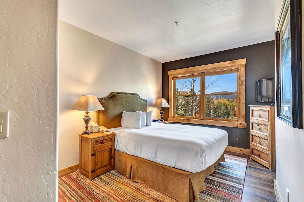 Un dormitorio con una cama grande, cabecera de madera, dos mesitas de noche con lámparas, un aparador y una ventana con vista a árboles.