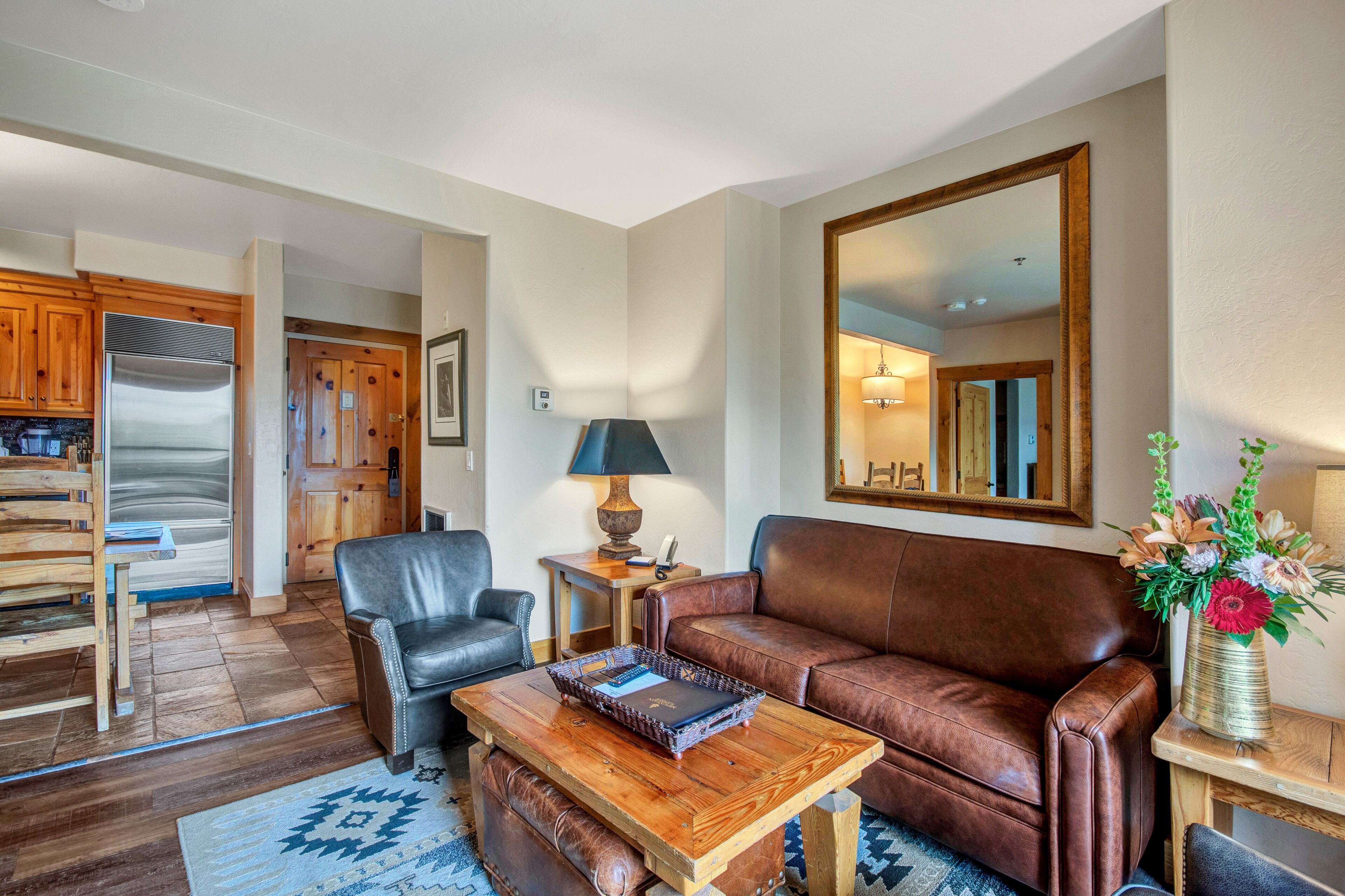 Un salon avec un canapé en cuir marron, une table basse en bois sur laquelle repose un livre, un fauteuil en cuir noir, une table d’appoint en bois avec une lampe, et un grand miroir accroché au mur.