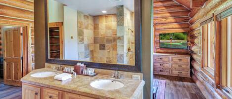 A log cabin bathroom with two sinks, a large mirror, wooden cabinets, and a view of a forest through a window.