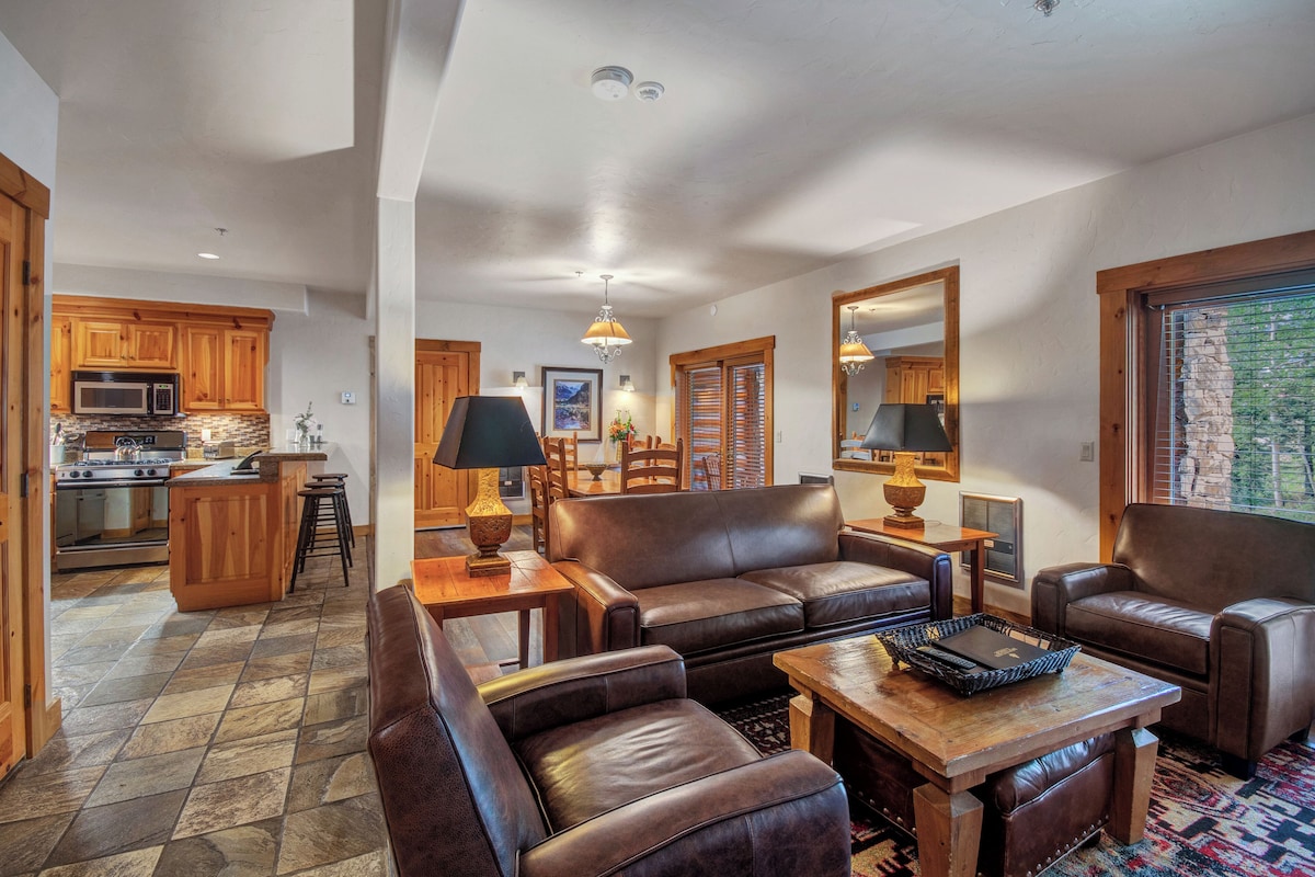 A spacious living area with a fireplace, leather sofas, a coffee table, and a kitchen in the background.