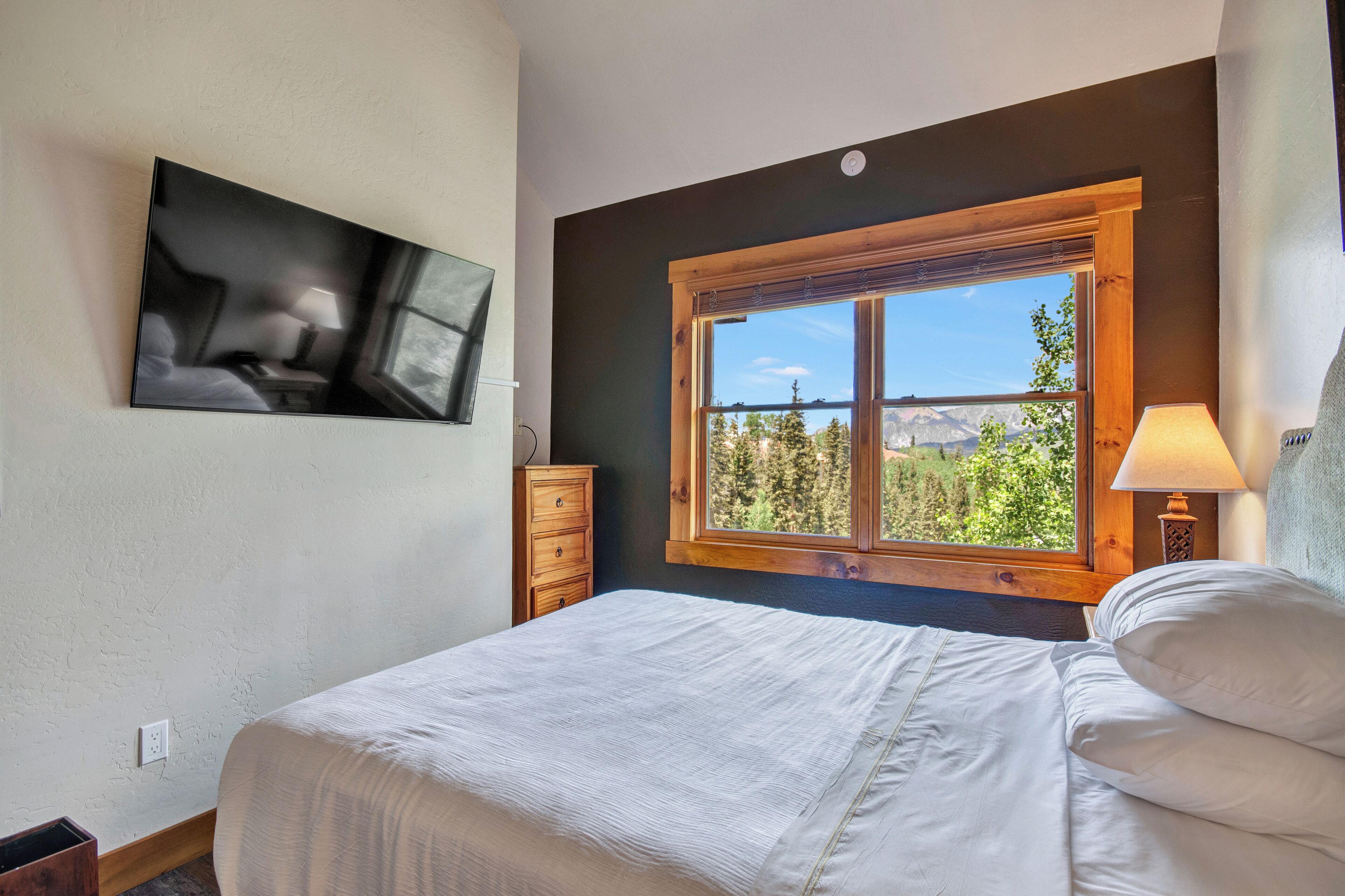 A bedroom with a bed, a nightstand, a lamp, a dresser, and a window with a view of trees and mountains.