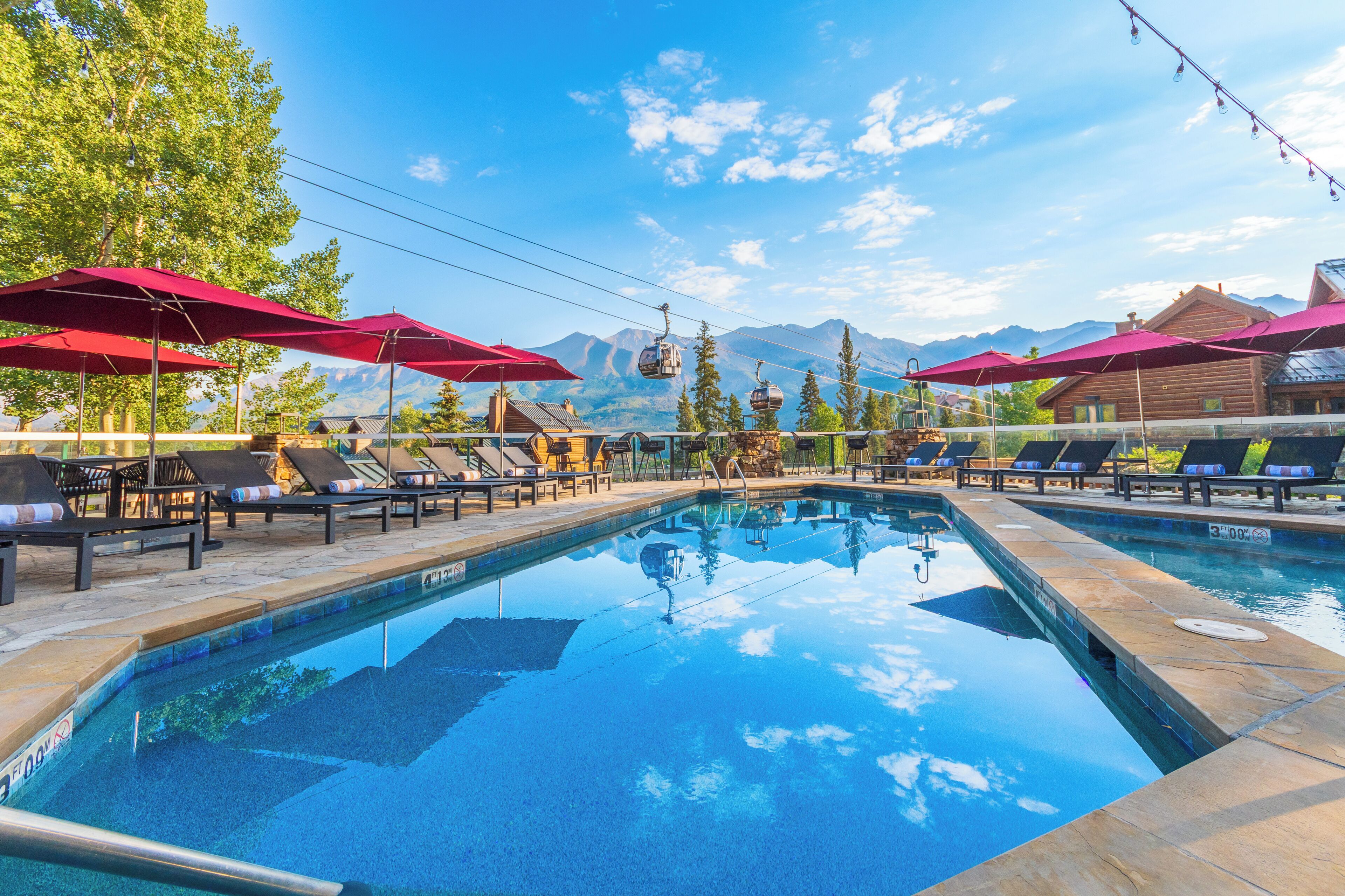 A pool area with lounge chairs and umbrellas, a cable car in the background, and mountains in the distance.
