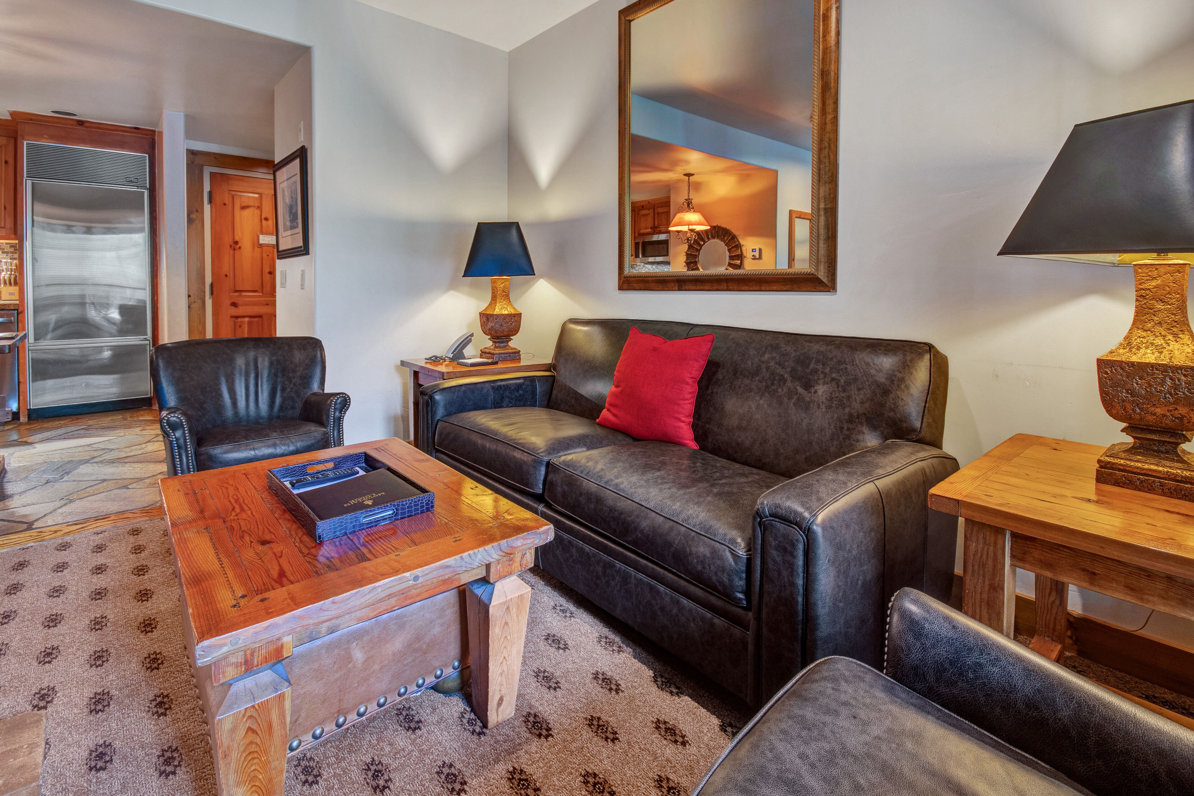 A living room with a leather sofa, a wooden coffee table, and a matching armchair. A large mirror hangs on the wall, and a lamp is on a side table.