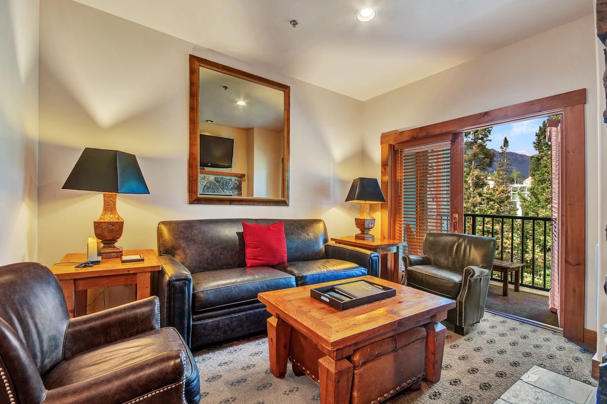 Una sala de estar con sofá, dos sillones, una mesa de centro y televisor. Hay un balcón con vista a árboles y montañas.