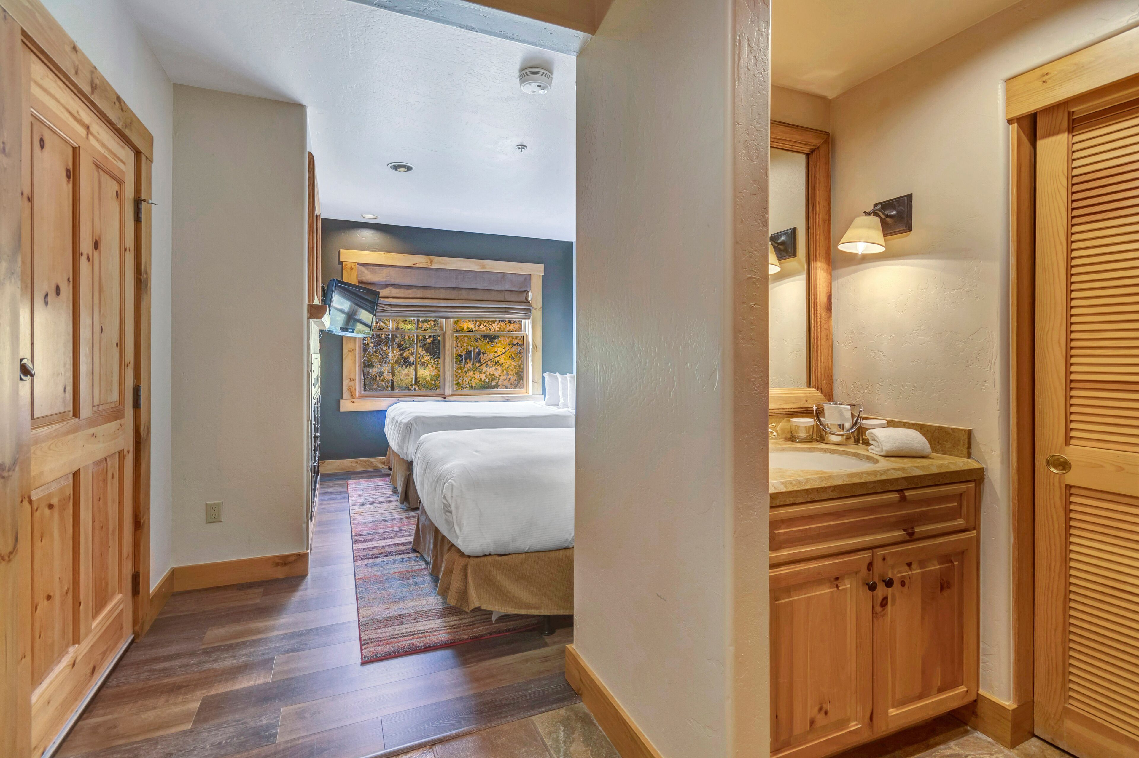 A hallway with wooden flooring, a bed with white bedding, a window with a view, and a bathroom with a sink and mirror.