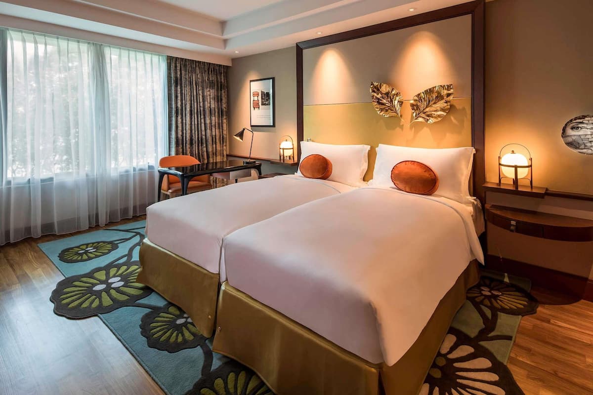A hotel room with two beds, a desk, a chair, a desk lamp, a wall-mounted lamp, and a large mirror.