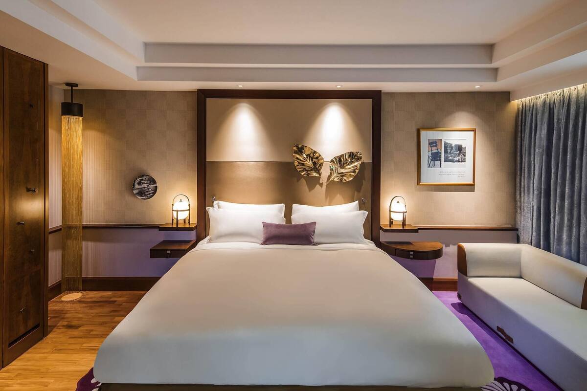 A hotel room with a large bed, a sofa, a nightstand with a lamp, a wall-mounted artwork, and a door with a wooden panel.