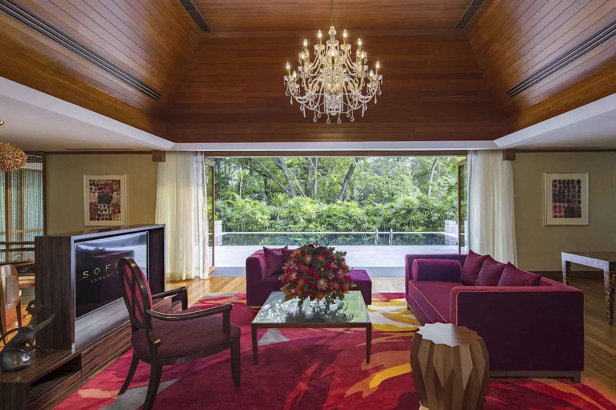 A spacious living room with a large chandelier, a sofa set, a coffee table with a floral centerpiece, and a television on a wooden stand.