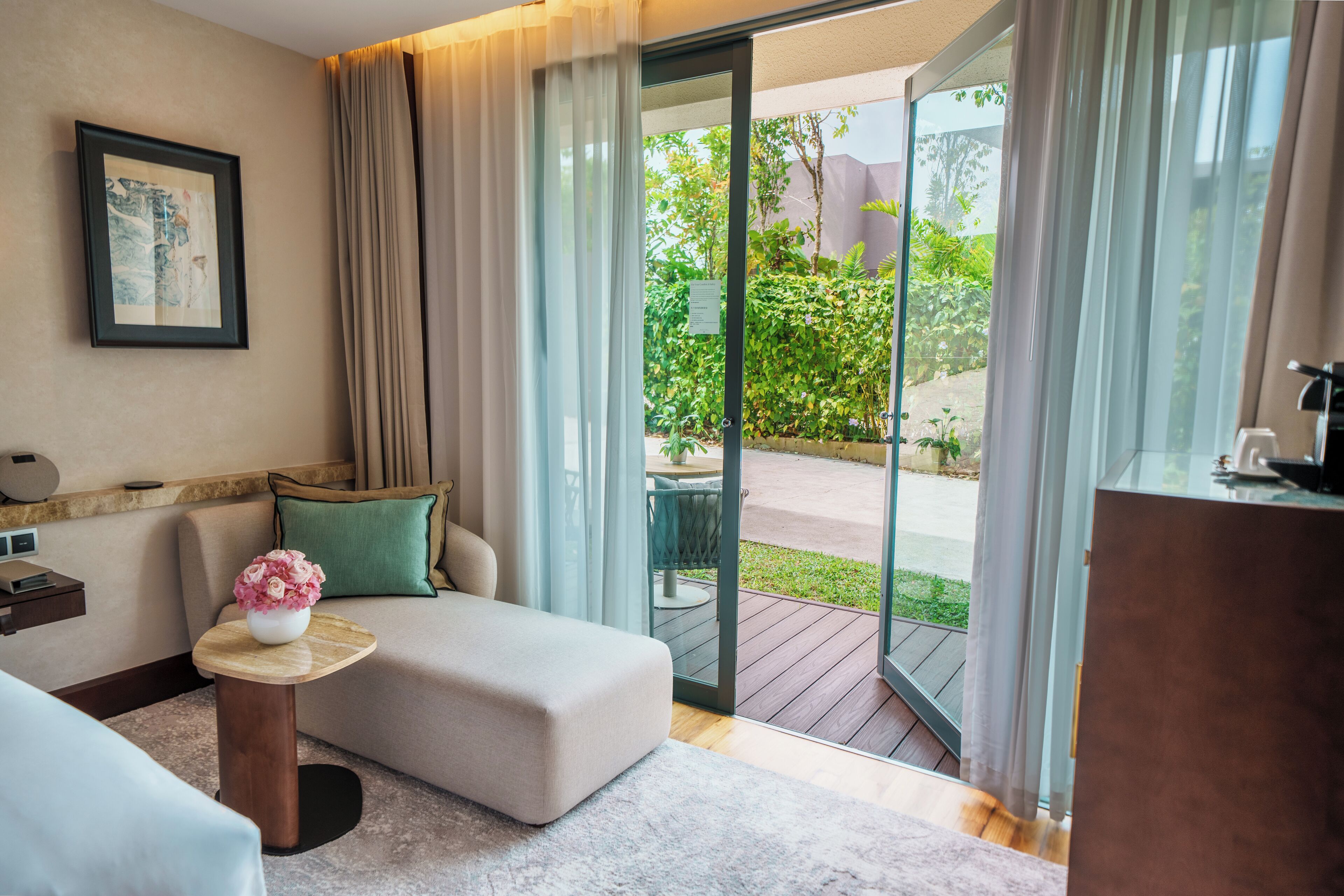 A modern hotel room with a sofa, a small round table with a vase of flowers, and a view of a garden through a sliding glass door.