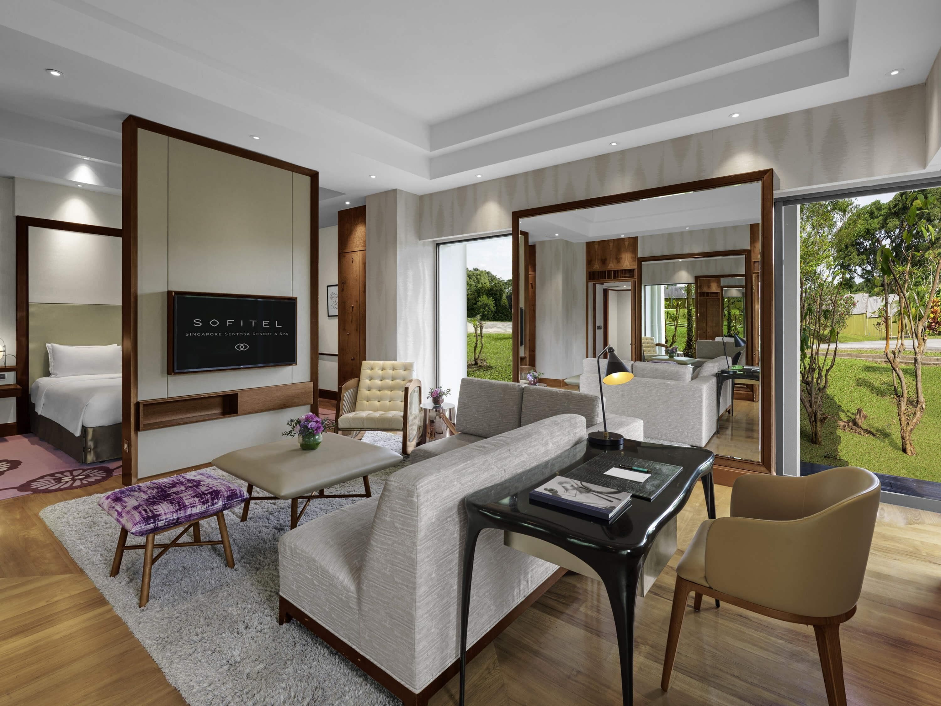 A modern hotel room with a sofa, armchair, and a TV on the wall.