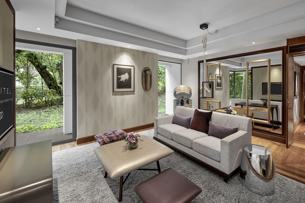 A spacious living room with a large sofa, a coffee table, and a TV. There is a large mirror reflecting the room and a view of the garden outside.