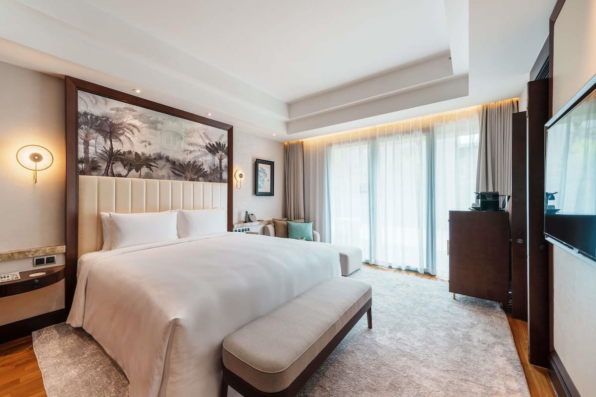 A large bed with a headboard, a bench, a desk, a television, and a painting on the wall.