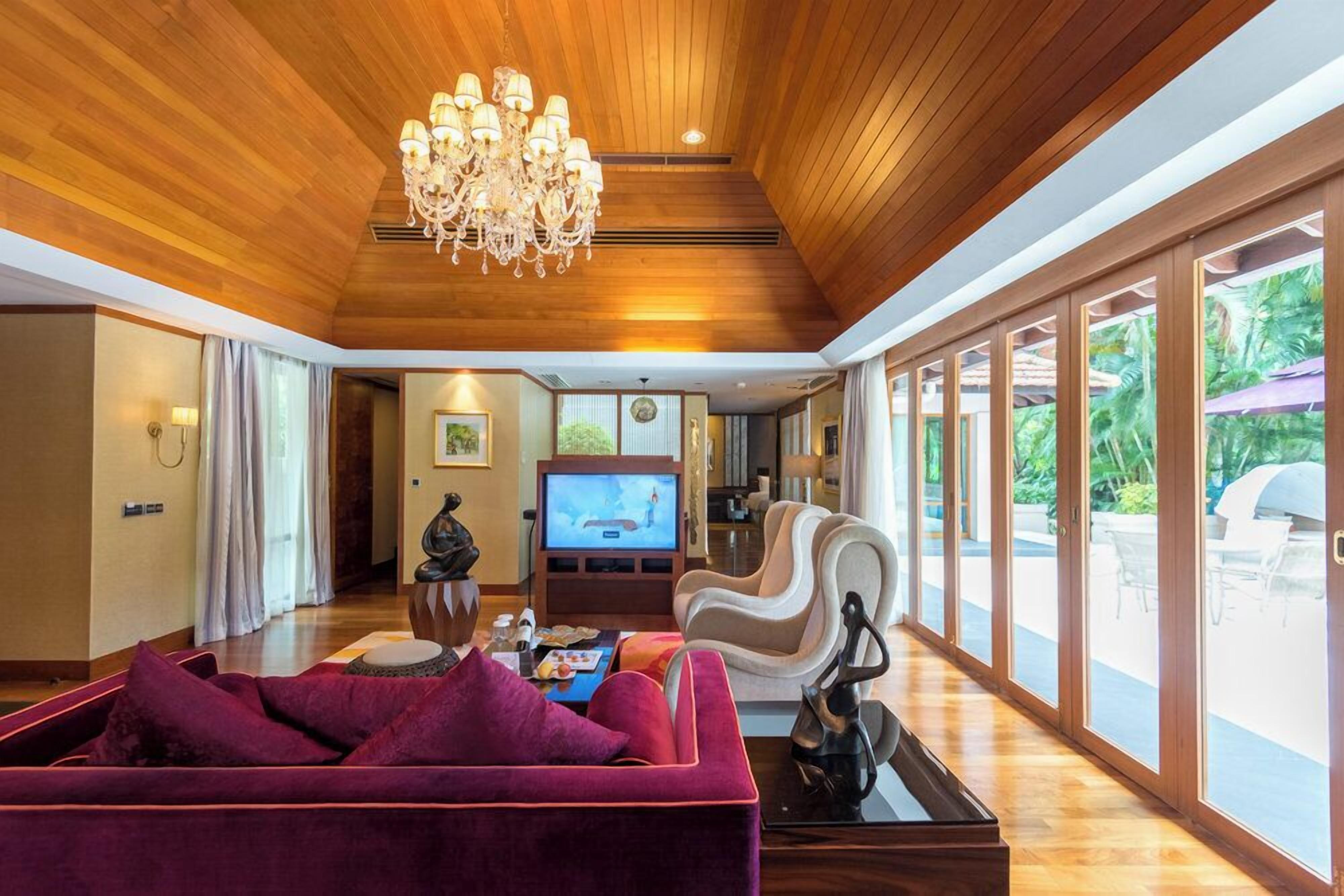 Un salón espacioso con techo de madera, una araña, televisor y un gran ventanal con vistas a un jardín.