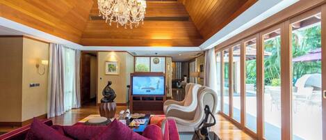 Un salón espacioso con techo de madera, una araña, televisor y un gran ventanal con vistas a un jardín.