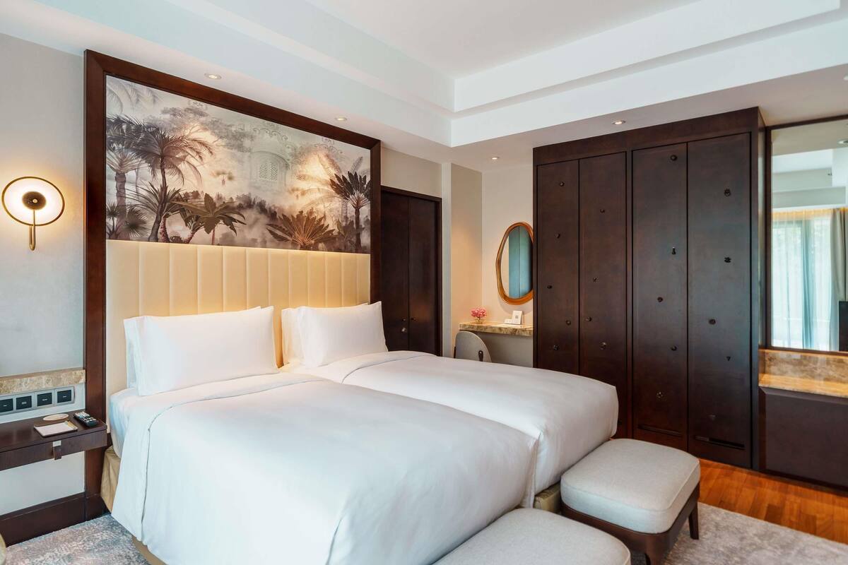 A hotel room with a large bed, a desk with a lamp, a painting on the wall, a wardrobe, and a mirror.
