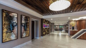 A modern hotel lobby with wooden panel ceilings, a central staircase, and large abstract paintings on the walls.