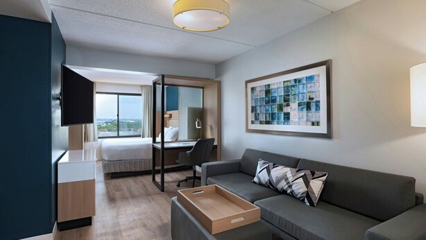 Ein modernes Hotelzimmer mit einem Bett, einem Schreibtisch, einem grauen Sofa, einem Fernseher und einem großen Fenster mit Blick auf die Stadt.