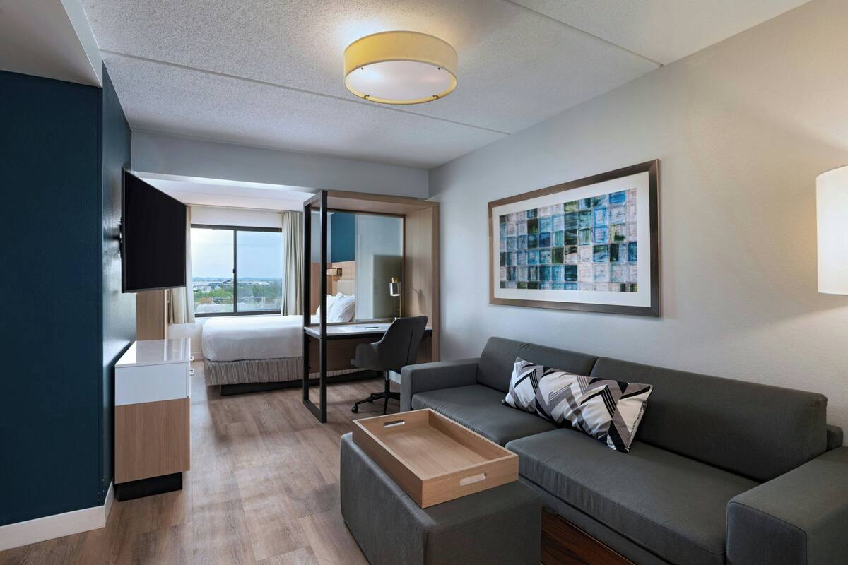 Ein modernes Hotelzimmer mit einem Bett, einem Schreibtisch, einem grauen Sofa, einem Fernseher und einem großen Fenster mit Blick auf die Stadt.
