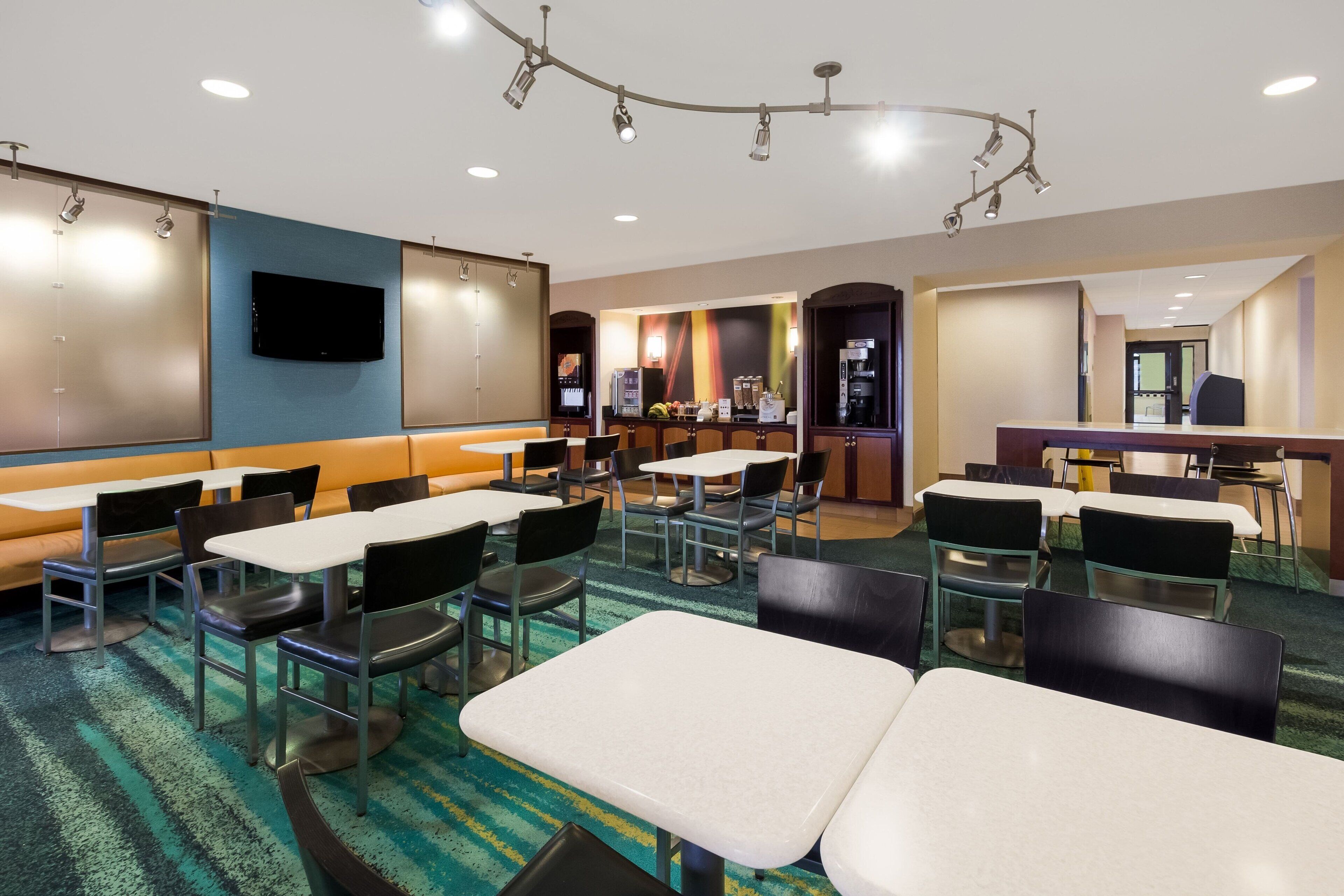 Une salle à manger d’hôtel avec plusieurs tables et chaises, un téléviseur fixé au mur et un appareil à café en arrière-plan.