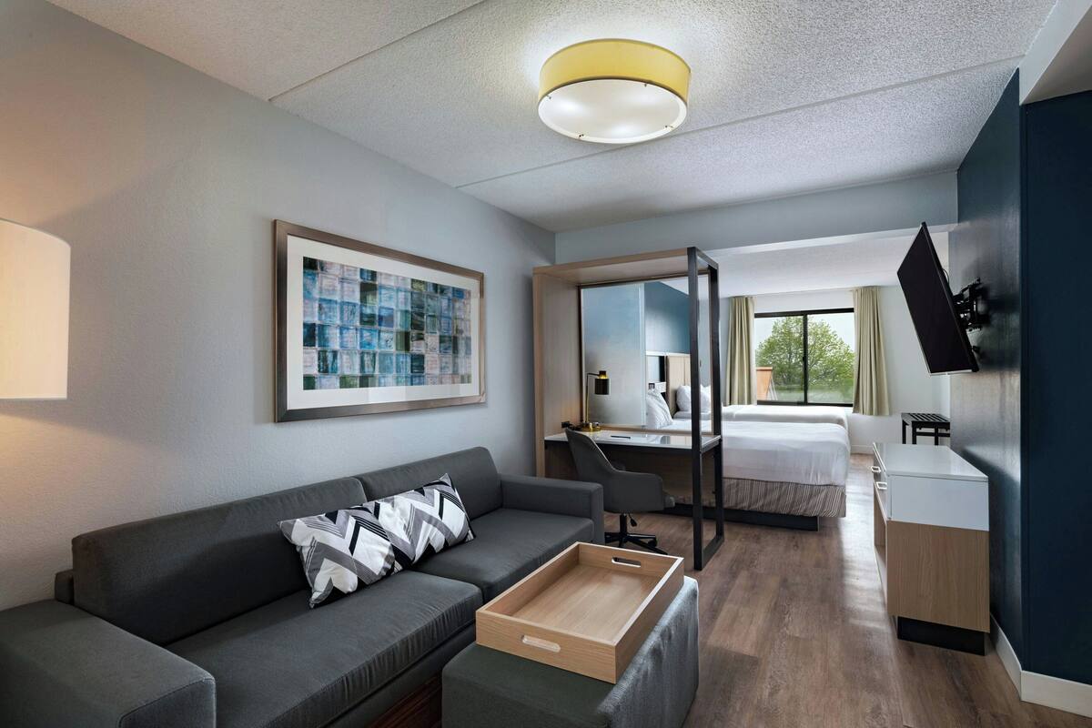 Ein modernes Hotelzimmer mit grauem Sofa, einem Holztisch, einem Schreibtisch, einem Bett und einem an der Wand montierten Fernseher.