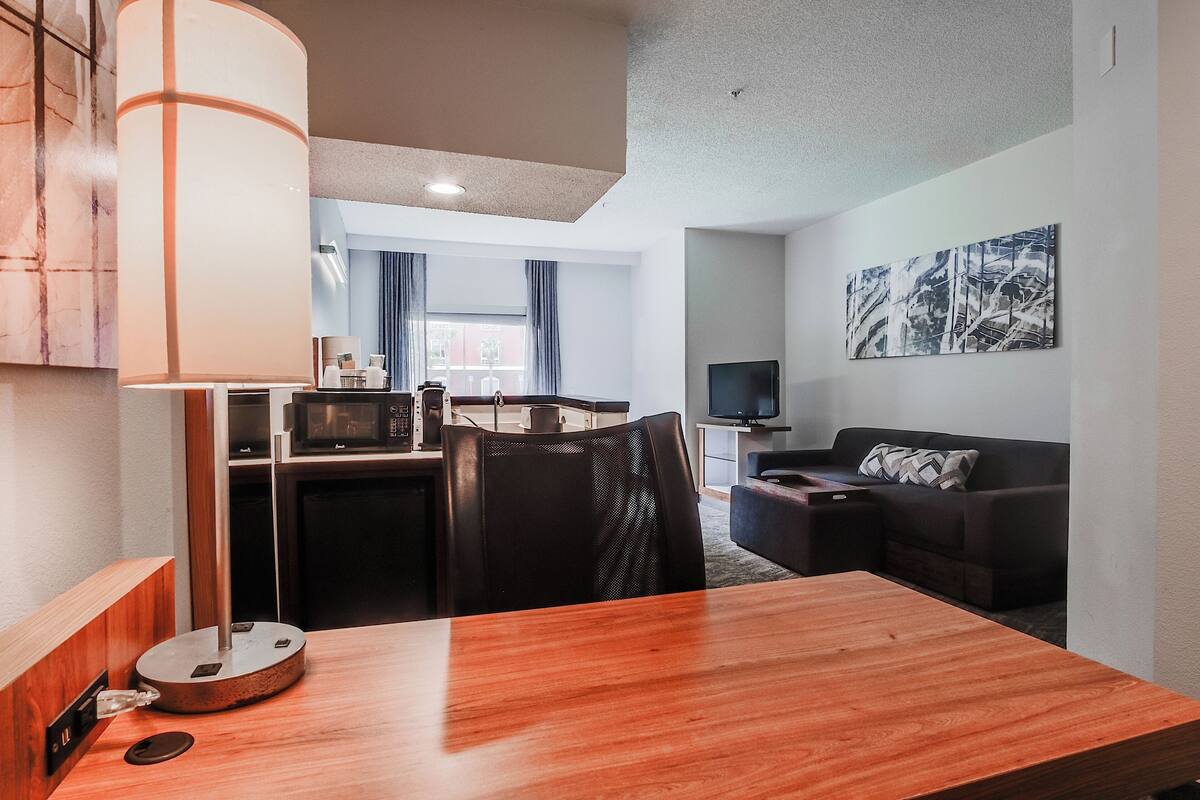 A modern hotel room with a wooden dining table, a black office chair, a desk with a computer, a flat-screen TV, a sofa, and a large abstract painting on the wall.
