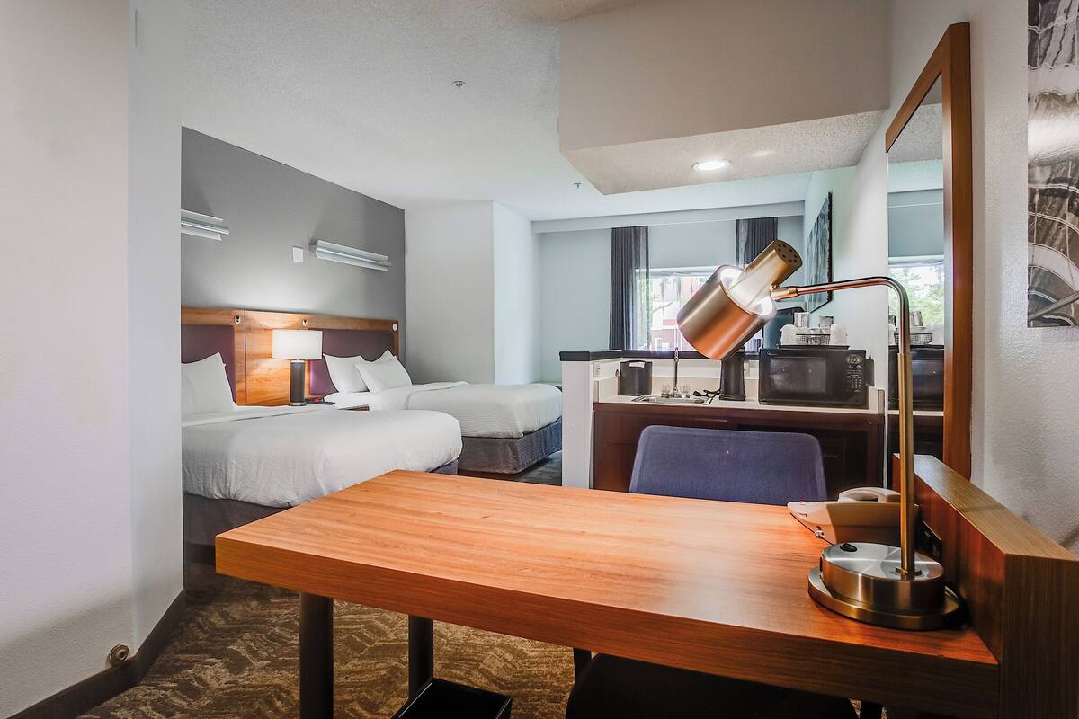 A hotel room with a wooden desk, a lamp, a bed, a chair, and a kitchenette with a microwave.