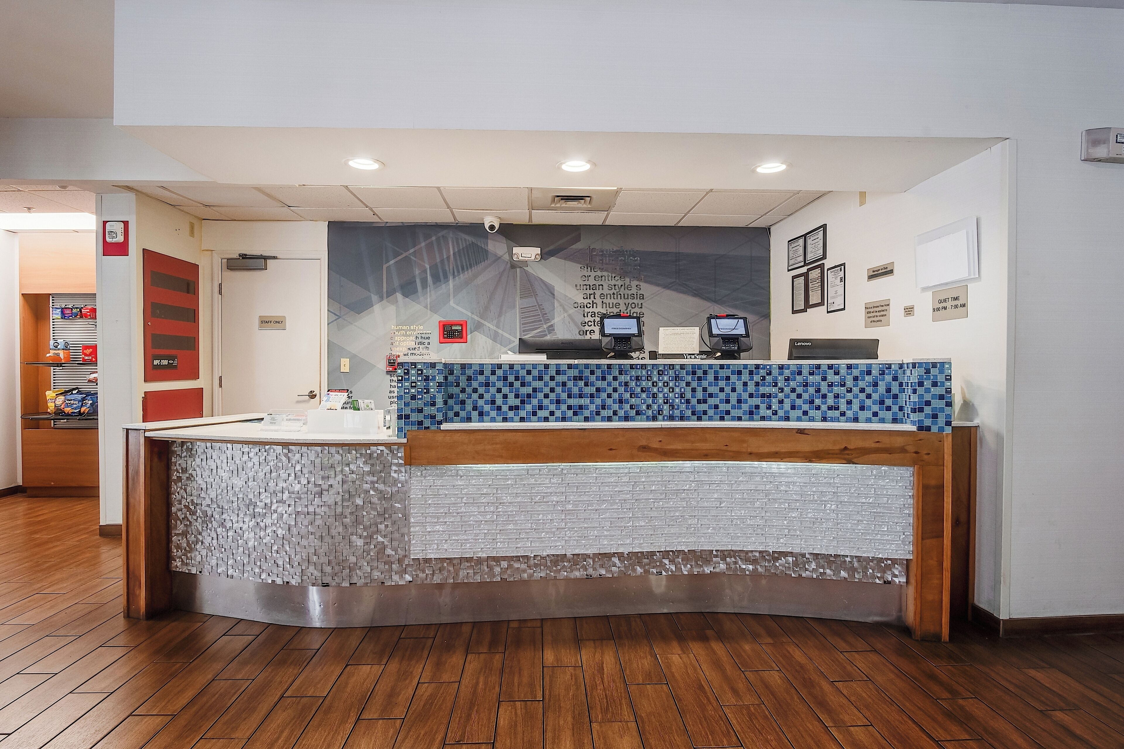 Un comptoir d’accueil avec un plateau supérieur en mosaïque bleue, un comptoir en bois et une base blanche. On y trouve deux téléphones, une enseigne au-dessus du comptoir, ainsi qu’un mur orné de tableaux et d’une horloge.