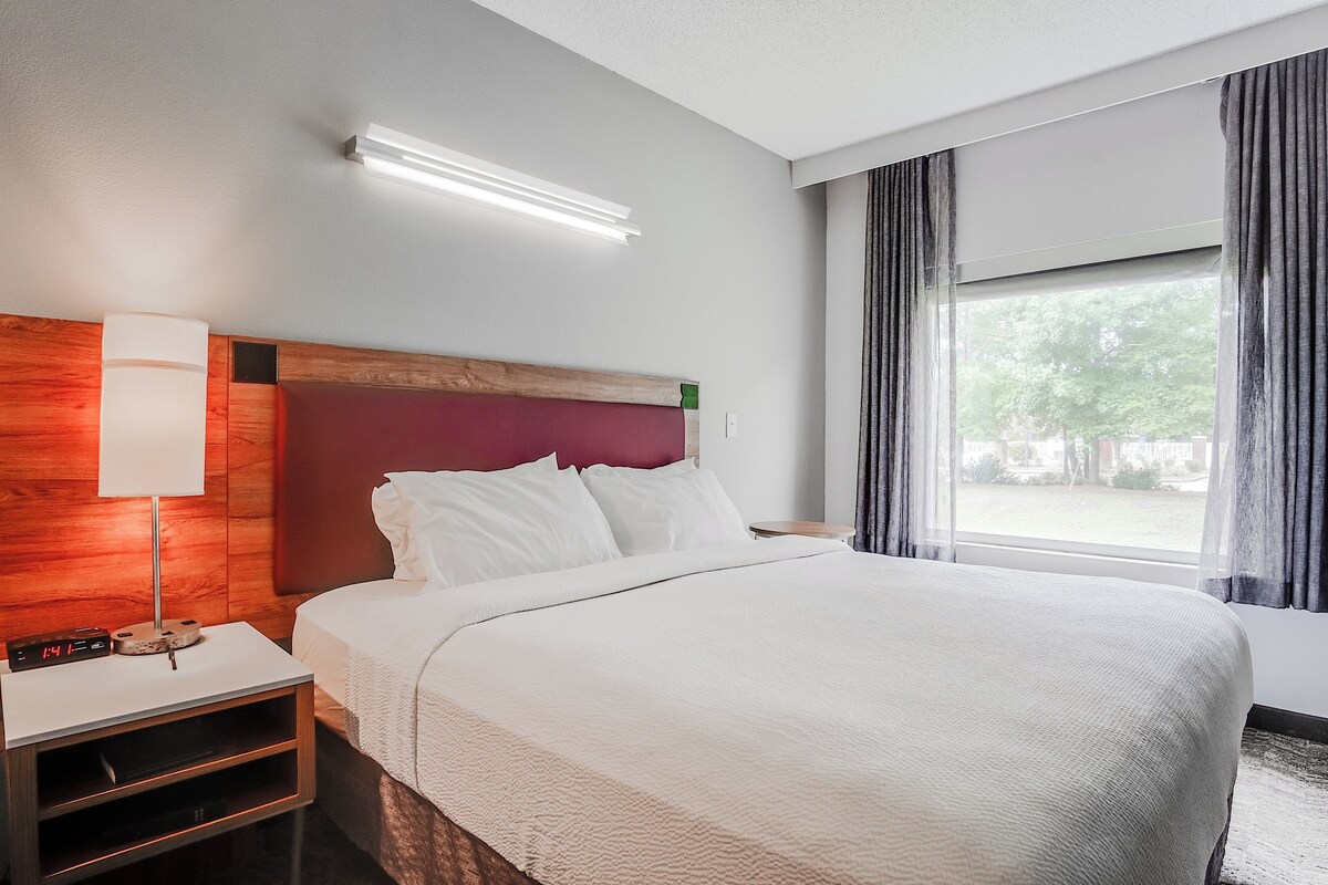 A hotel room with a large bed, a nightstand with a lamp, a digital clock, and a window with curtains.