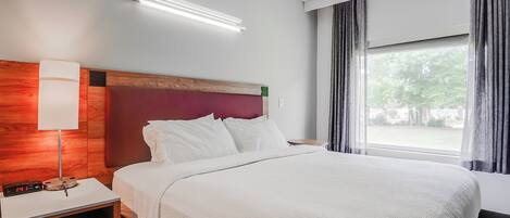 Una habitación de hotel con una cama grande, una mesita de noche con lámpara, un reloj digital y una ventana con cortinas.
