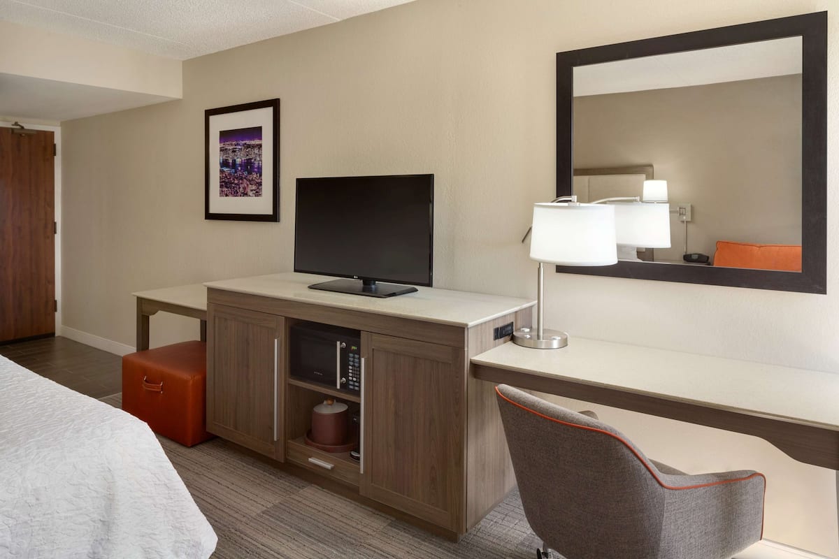 A hotel room with a bed, a desk with a flat-screen TV, a lamp, a chair, and a cabinet with a microwave.