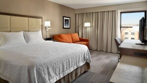 Ett hotellrum med en stor säng, ett skrivbord, en stol och utsikt över byggnader genom fönstret.