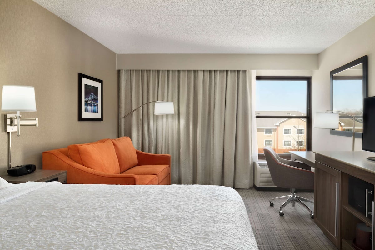 A hotel room with a large bed, an orange armchair, a desk with a chair, a large window with a view of buildings, and a framed picture on the wall.