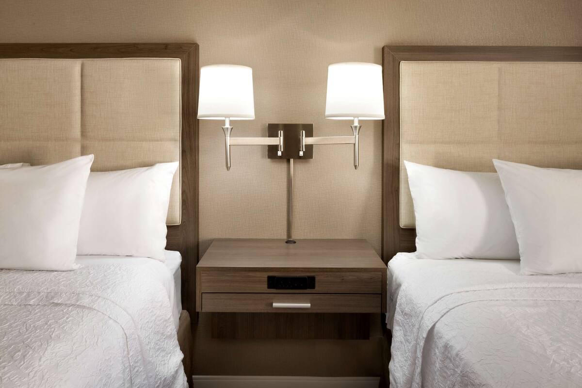 A hotel room with two beds, each with white linens and pillows, a wooden nightstand with a drawer, and two wall-mounted lamps.
