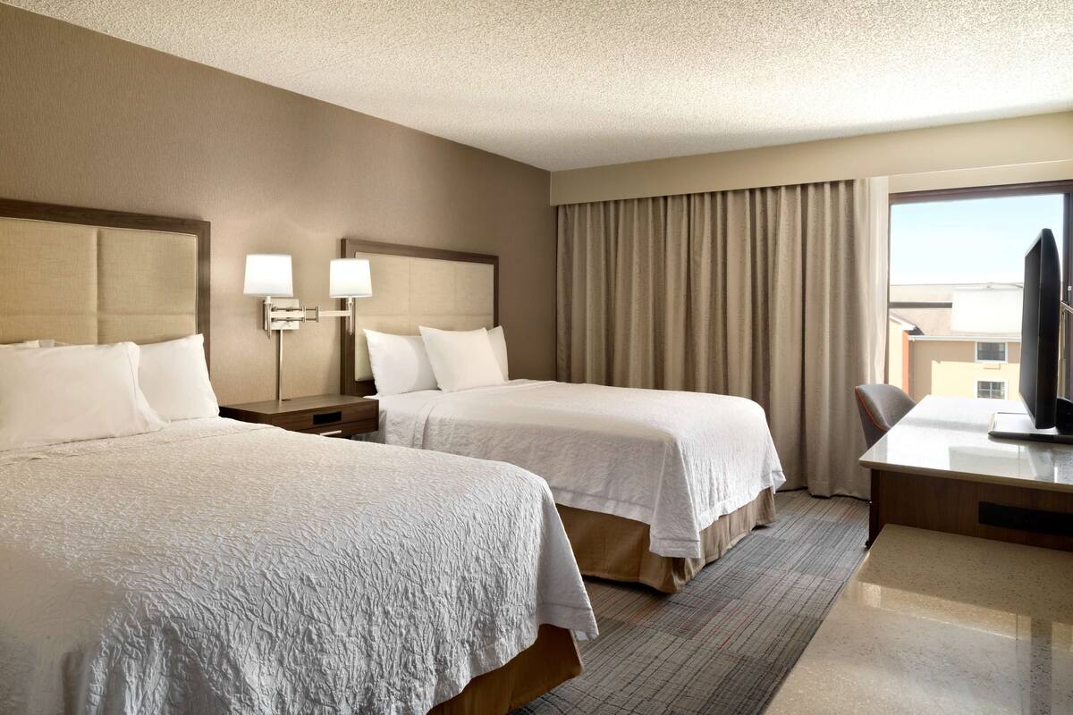 A hotel room with two beds, a desk, a TV, and a large window with curtains.