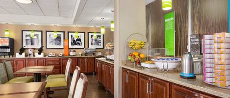 A hotel restaurant with wooden tables and chairs, a buffet counter with fruits and beverages, and framed pictures on the wall.