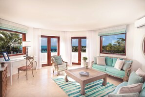 Uma sala de estar ampla com vista para o mar, uma secretária branca com cadeira, um sofá verde e uma mesa de centro.