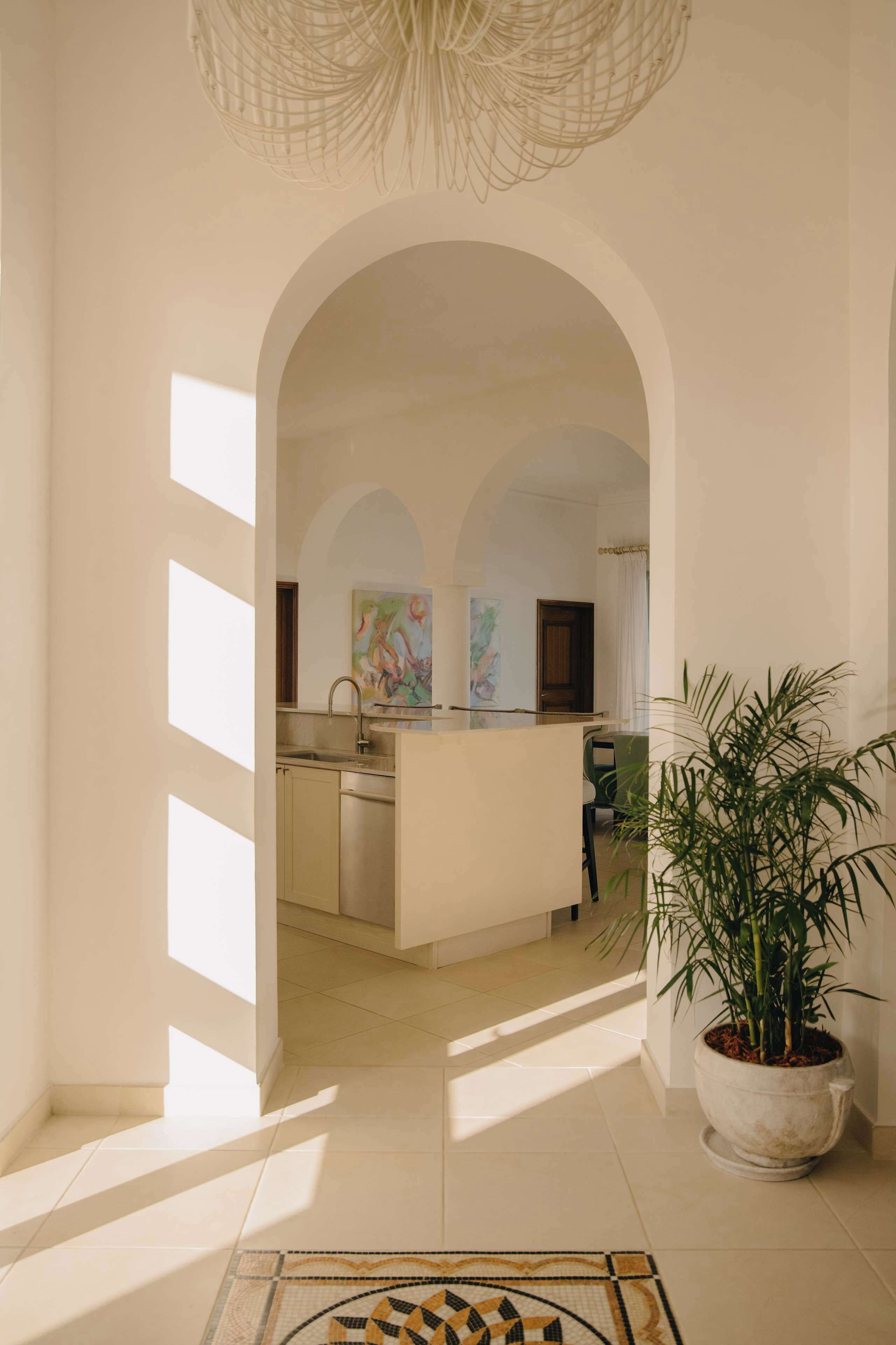 An interior view of a hotel lobby with a tiled floor, a large potted plant, and a kitchen area visible through an arched doorway.