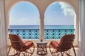 Varanda com duas cadeiras de vime e uma mesa pequena, com vista para um mar turquesa com um veleiro e uma linha costeira distante.