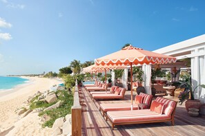 Un resort frente a la playa con sillones de descanso bajo sombrillas a rayas, una terraza de madera y vista al océano.