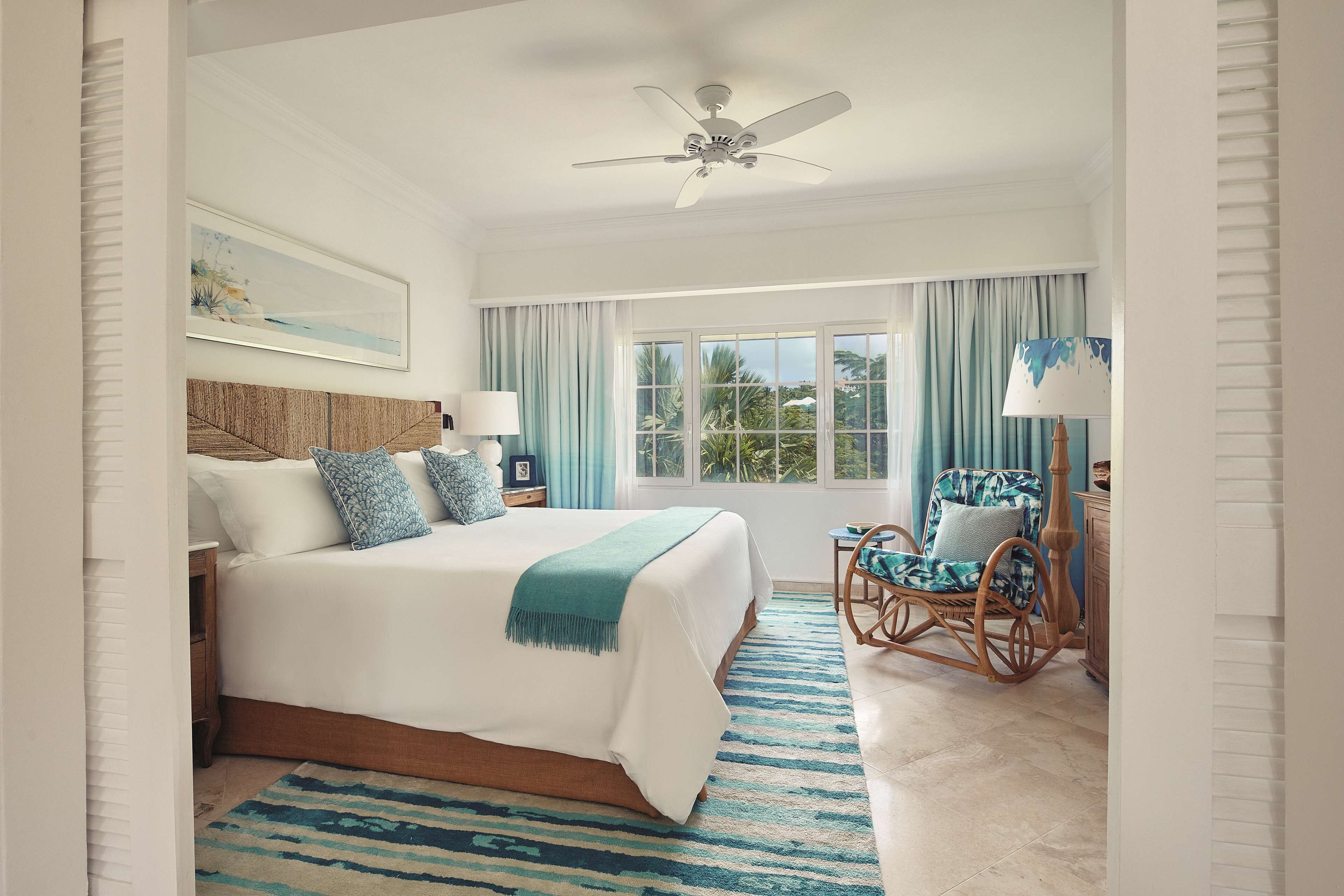 A bedroom with a large bed, a ceiling fan, a window with curtains, a rocking chair, and a nightstand with a lamp.