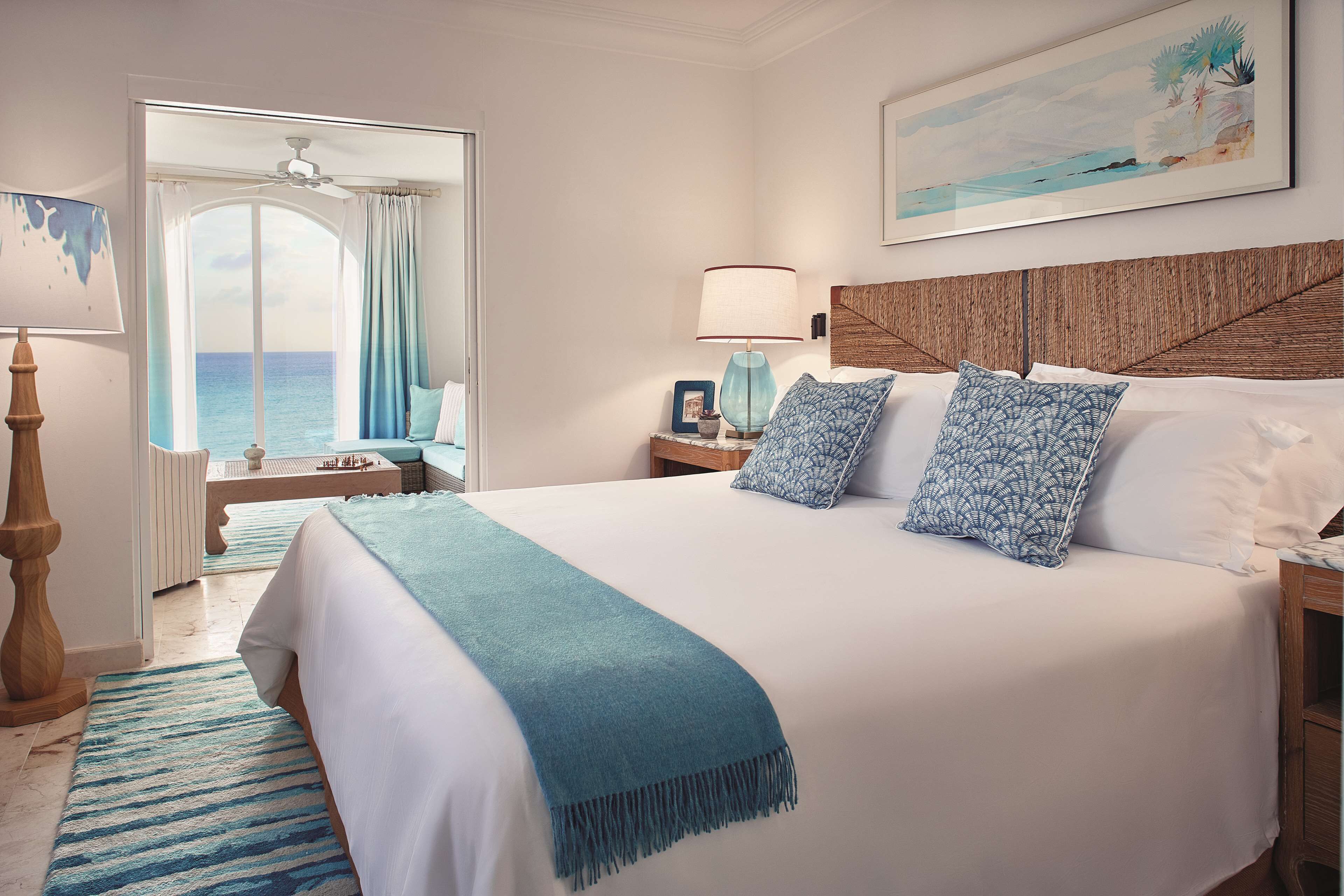 Une chambre avec un grand lit, une vue sur la mer, un ventilateur de plafond et un tableau encadré au mur.