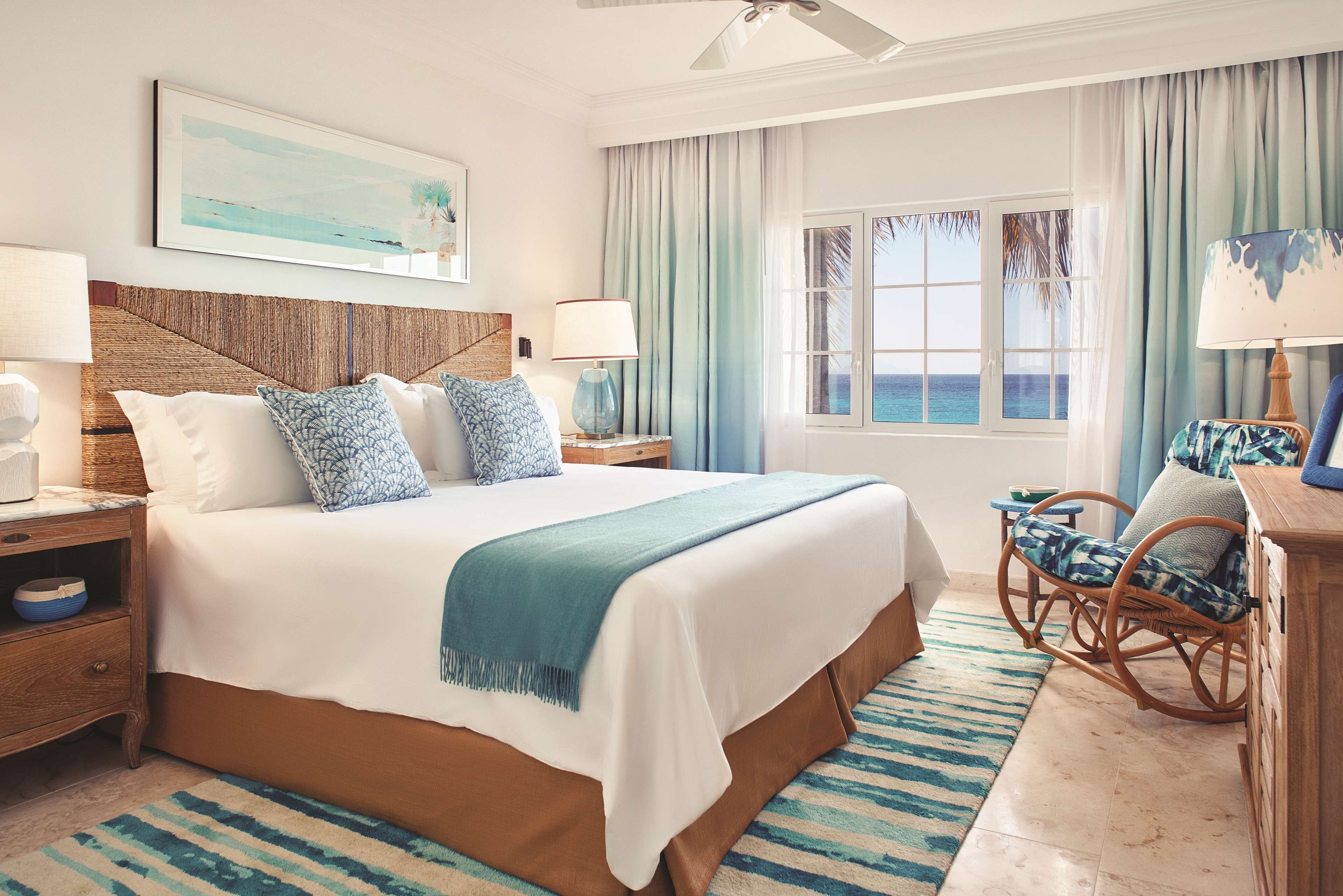 A hotel room with a large bed, two bedside tables, a chair, a view of the ocean, and a ceiling fan.